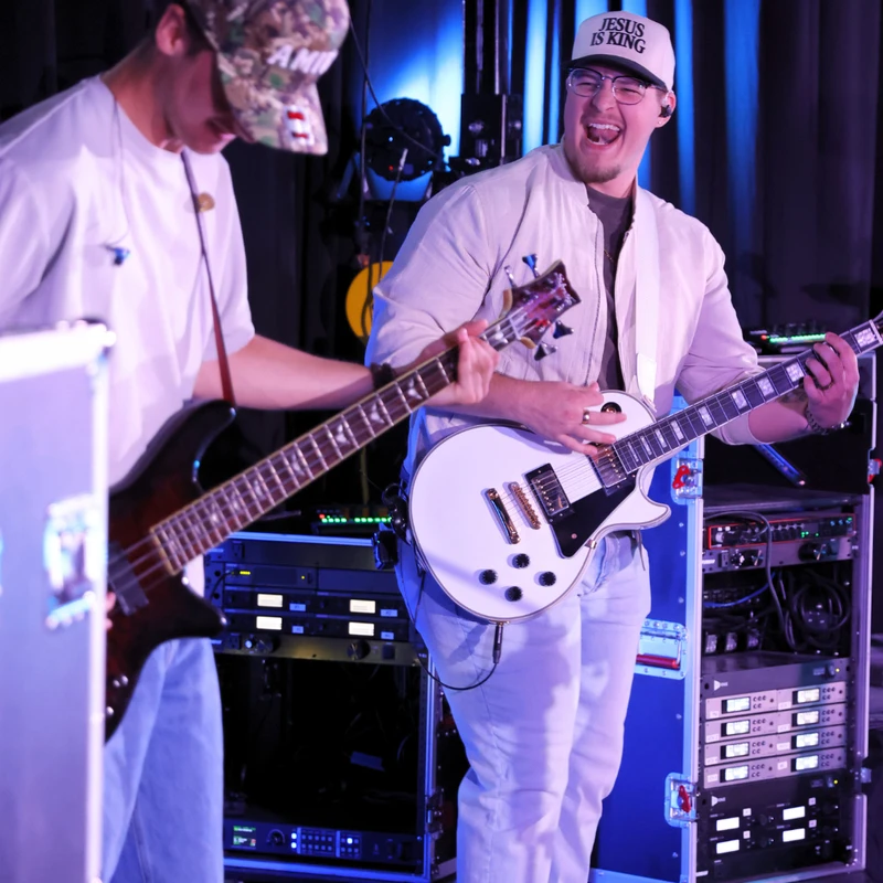 Two young men perform on a stage with blue lighting. One plays a bass guitar and the other smiles widely while playing a white electric guitar.