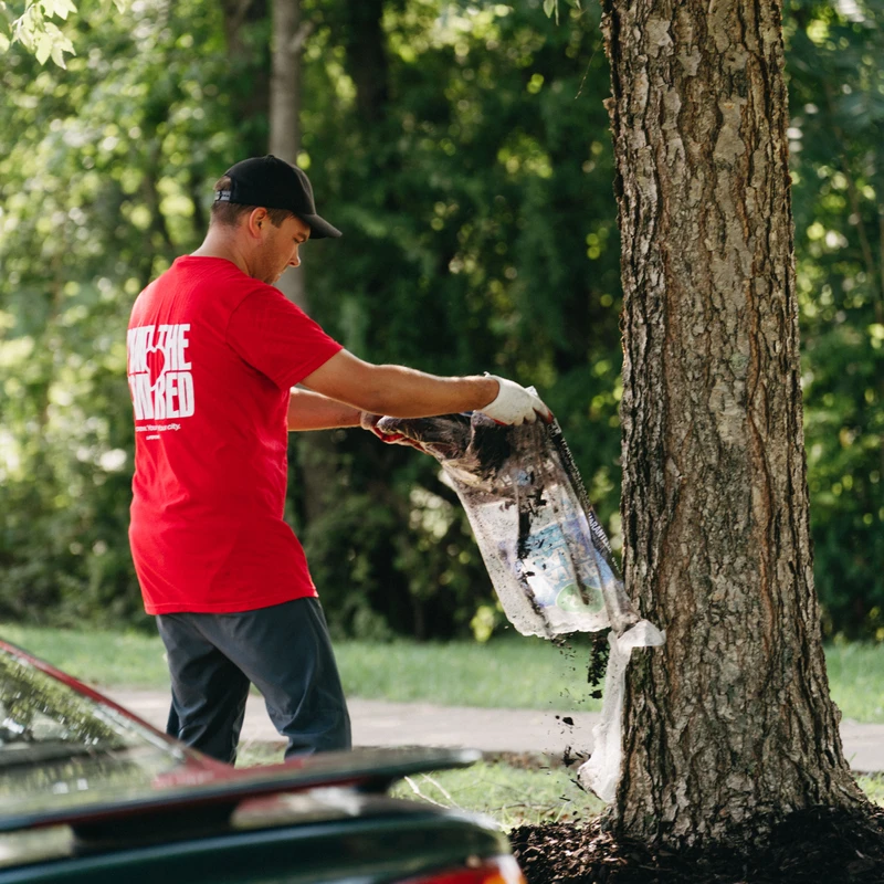 A man wearing a red t-shirt, black cap, and white gloves pours a bag of dark mulch around the base of a large tree on a sunny day.
