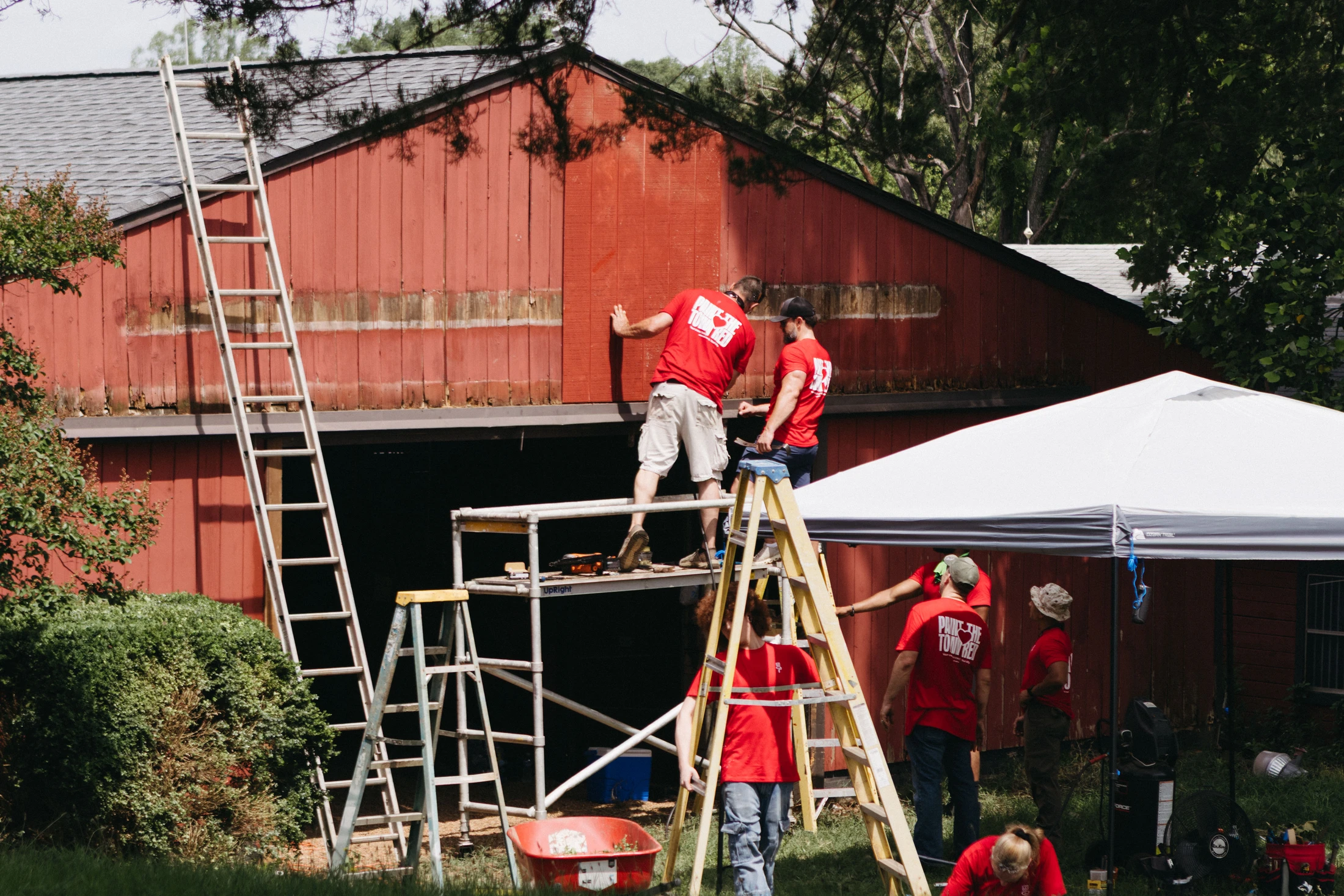 A group of volunteers in matching red shirts work together on a sunny day, using scaffolding and ladders to repair the siding of a large red barn.