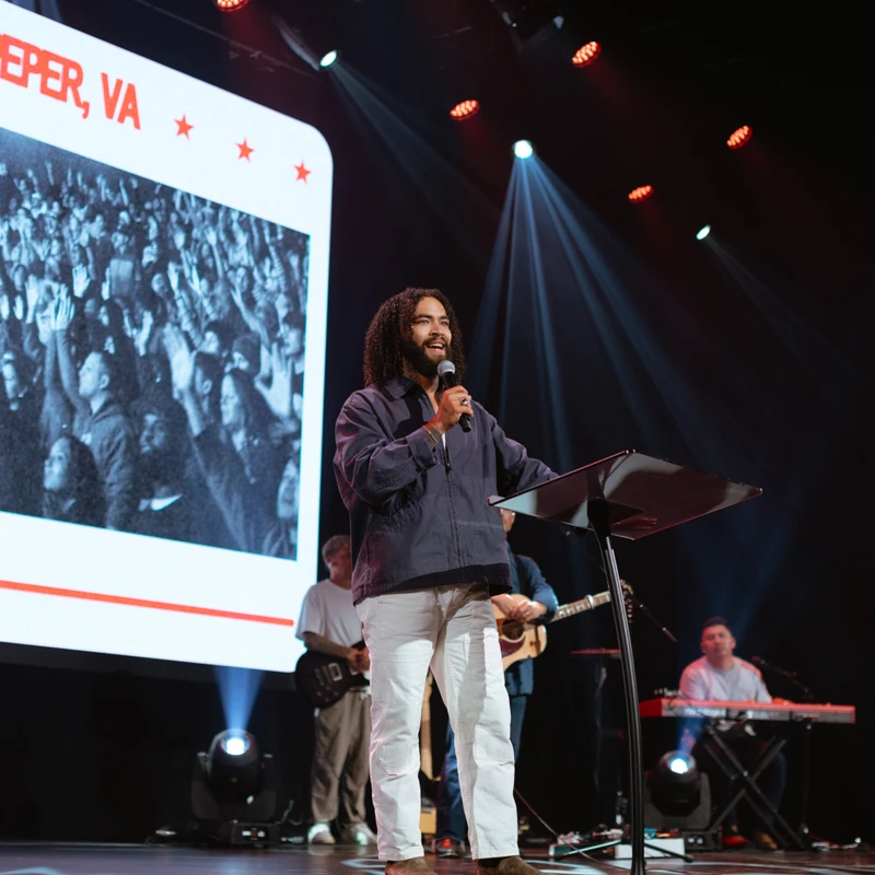 A man with long curly hair speaks into a microphone at a lectern on stage. A large screen behind him reads "CULPEPER, VA" above a photo of a crowd.