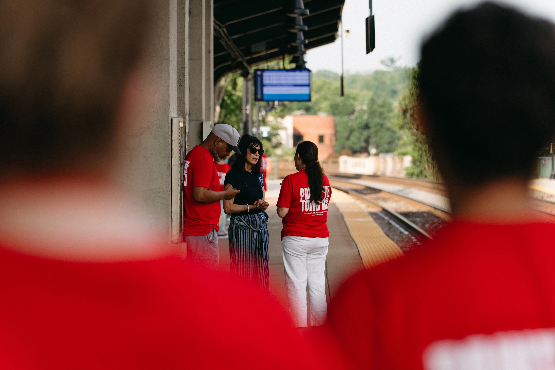 A group of people, some in matching red t-shirts, stand talking on an outdoor train station platform, framed by two blurred figures in the foreground.