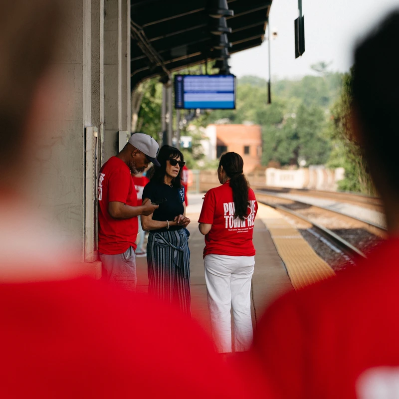 A group of people, some in matching red t-shirts, stand talking on an outdoor train station platform, framed by two blurred figures in the foreground.