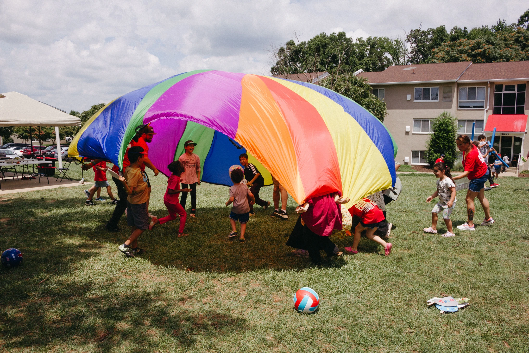 A group of children and a few adults play with a large, rainbow-colored parachute on a grassy lawn on a sunny day, with some kids running underneath it.