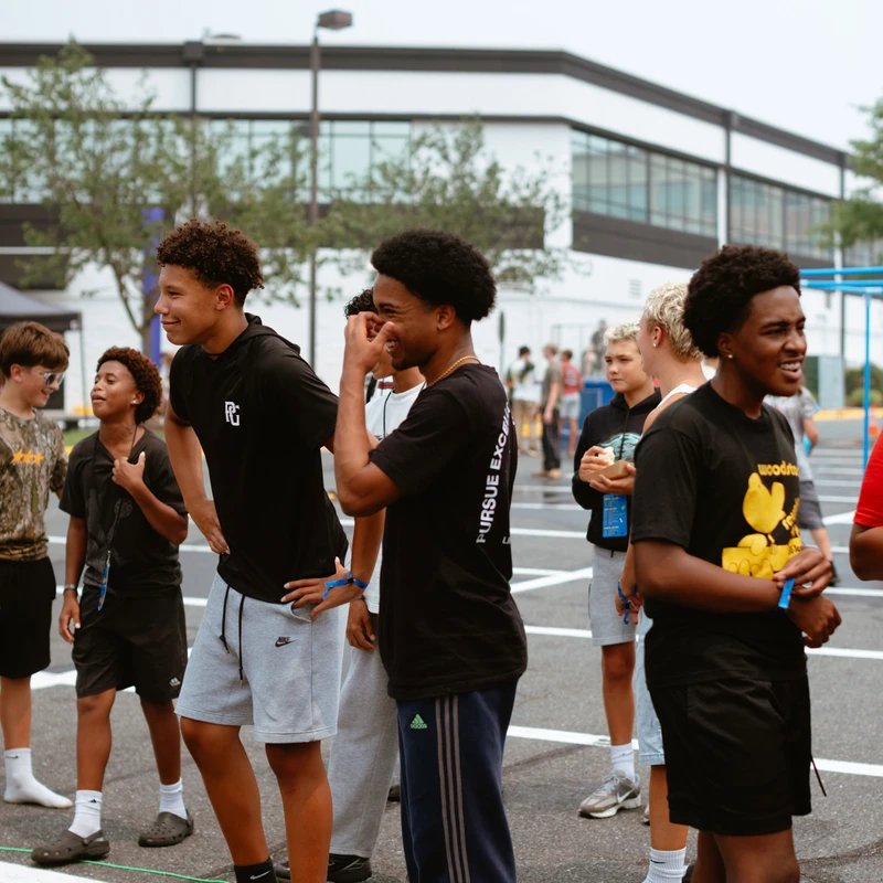 A diverse group of teenage boys in casual athletic clothing stand together in a parking lot, smiling and talking amongst themselves.