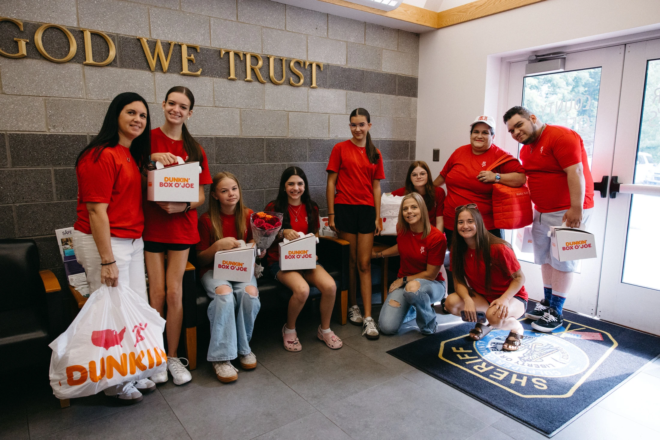 A group of volunteers in red shirts smiles for a photo while holding boxes of Dunkin' coffee and treats in a sheriff's office lobby.
