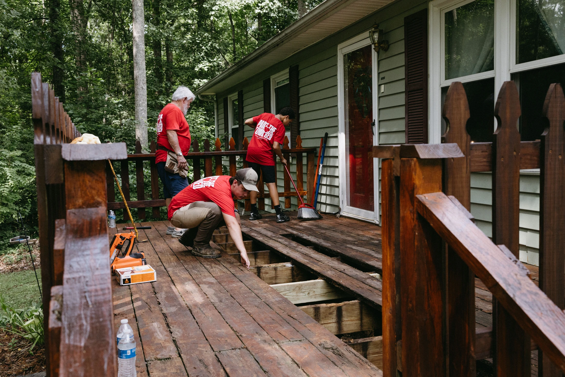 Three volunteers in matching red shirts work on repairing an old wooden deck. One kneels to fix the floorboards while another sweeps in the background.