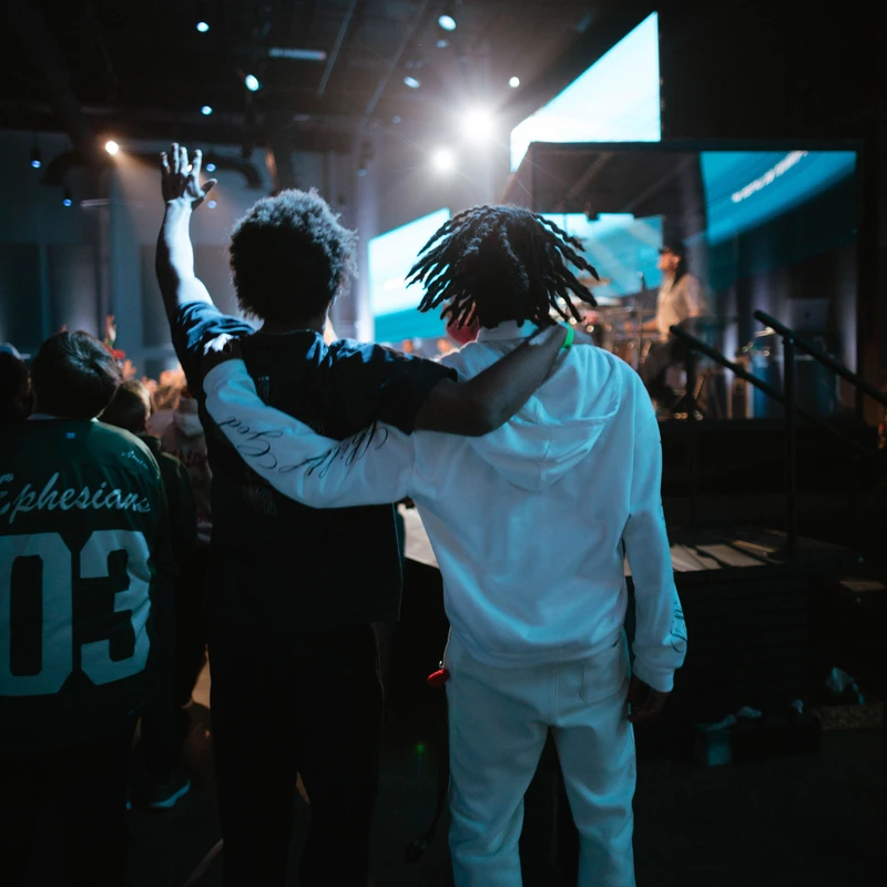 From behind, two young Black men stand in a crowd at a concert or church. One has his arm around his friend, who has one hand raised in the air.