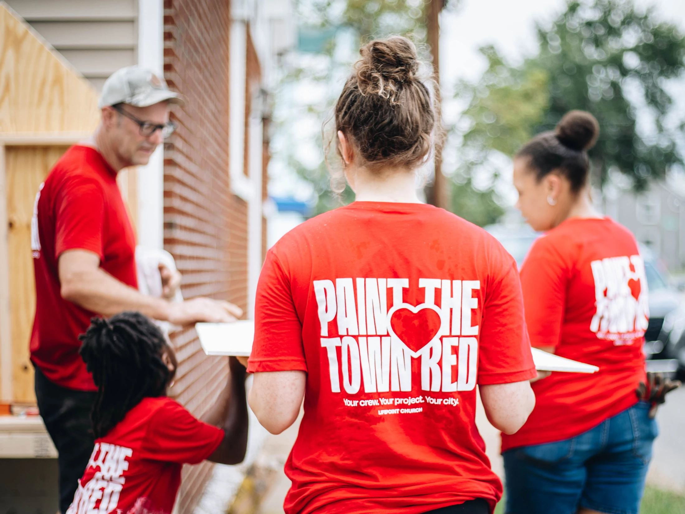A group of volunteers in matching red shirts that read 'Paint The Town Red' work on the side of a brick house during a community service event.