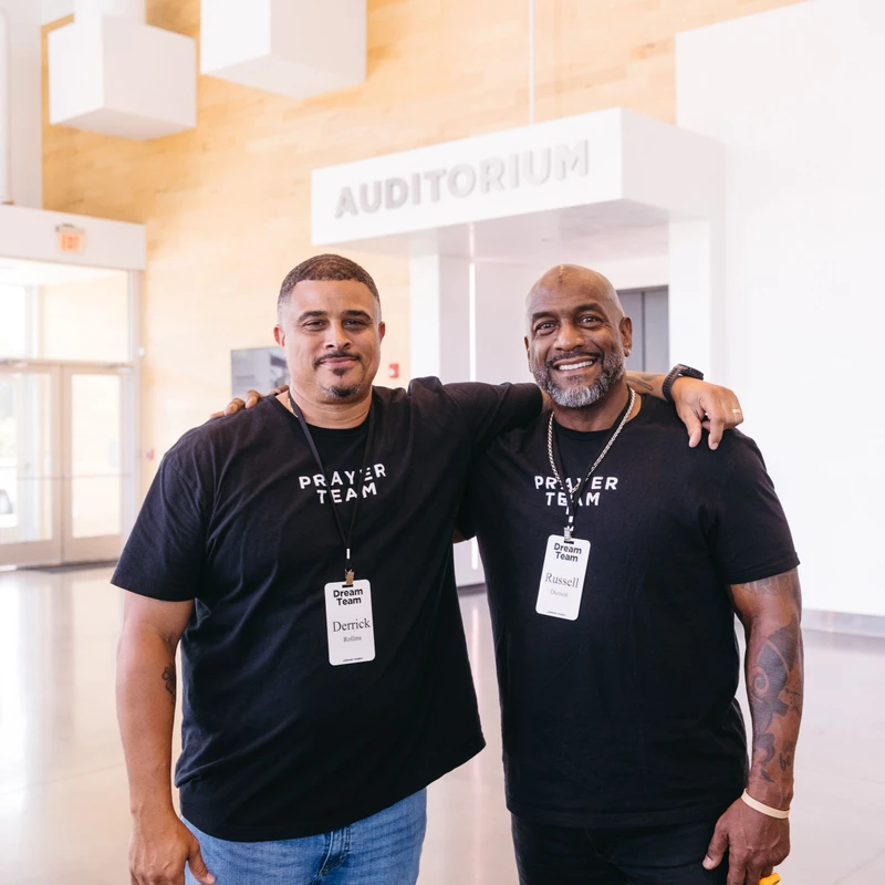 Two men in black 'Prayer Team' shirts stand with arms around each other, smiling in a bright lobby. A sign for an auditorium is visible in the background.