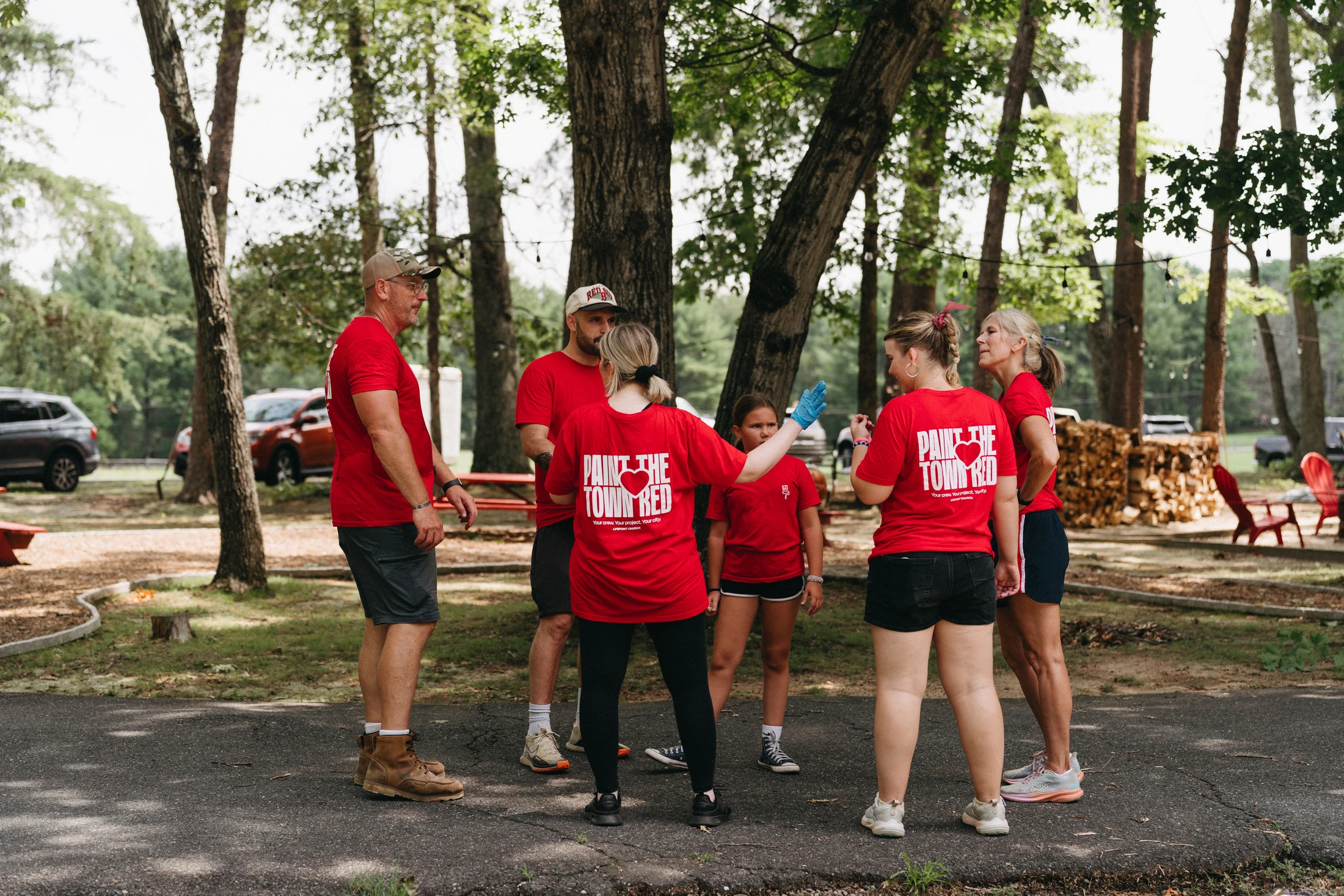 A group of volunteers wearing matching red 'Paint the Town Red' t-shirts stand in a circle outdoors, discussing plans for a community service event.