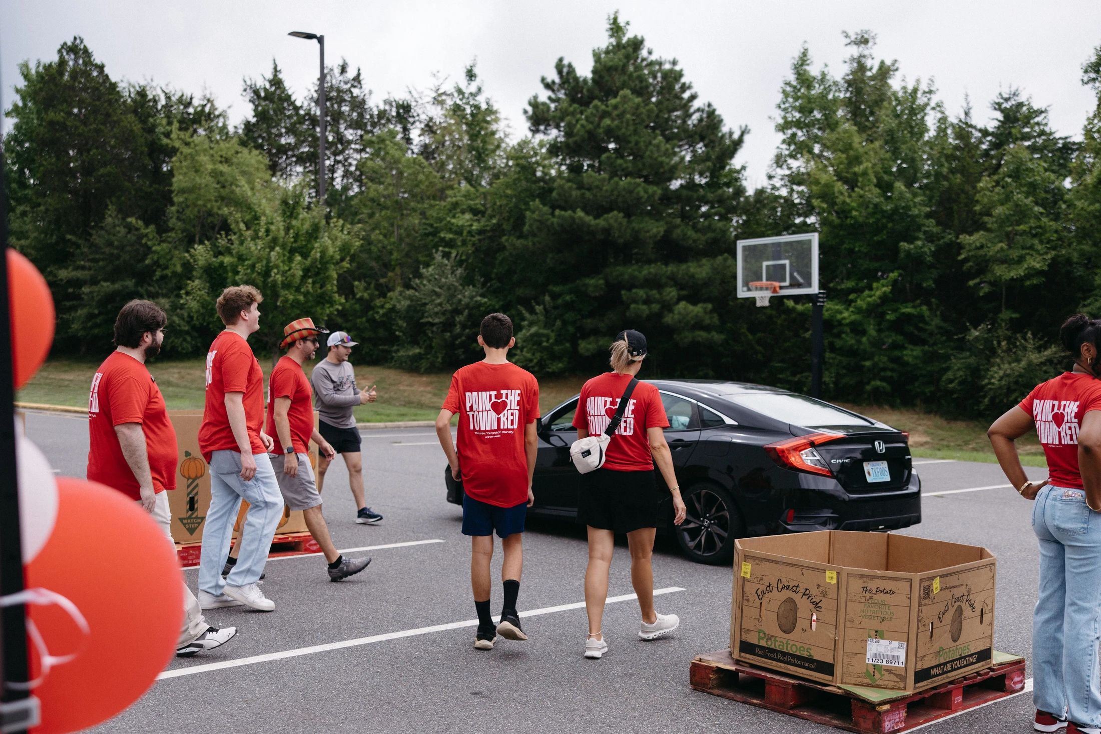 A group of volunteers in matching red "Paint the Town Red" t-shirts stand in a parking lot next to a box of potatoes during a community service event.