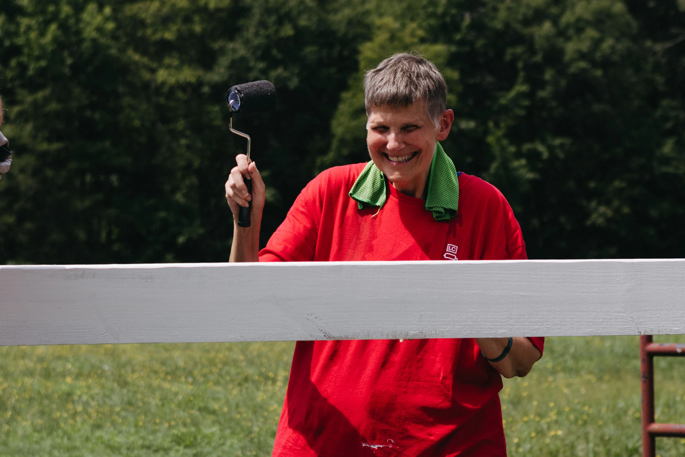 A smiling woman in a red t-shirt holds a small paint roller while painting a white wooden fence outdoors in a grassy field on a sunny day.