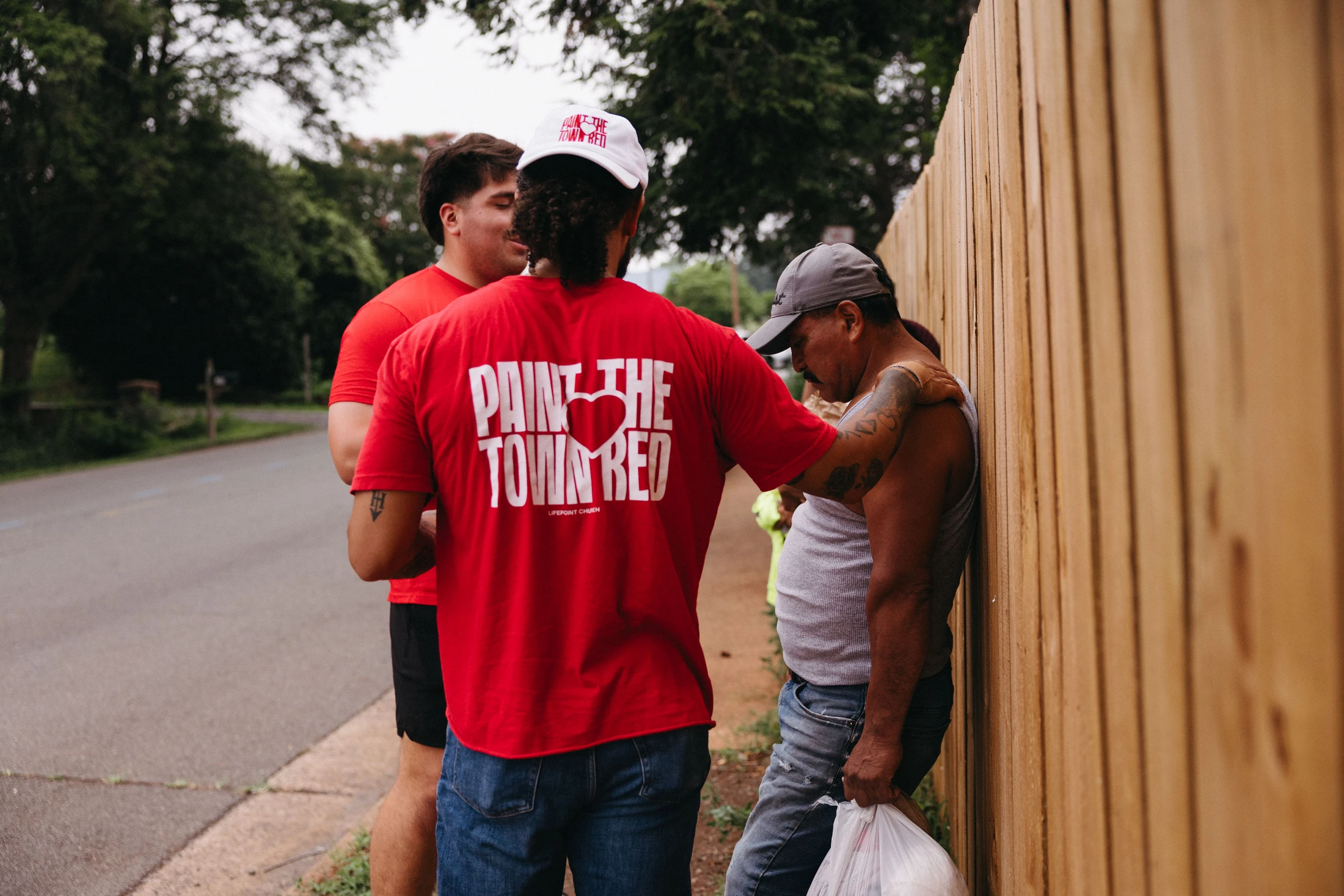 A volunteer in a red "Paint the Town Red" t-shirt places a comforting hand on another man who is leaning against a wooden fence on a sidewalk.