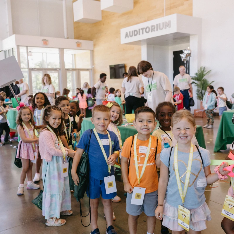 A diverse group of young children wearing name tags on lanyards smile happily for the camera while lining up for an event inside a large building.
