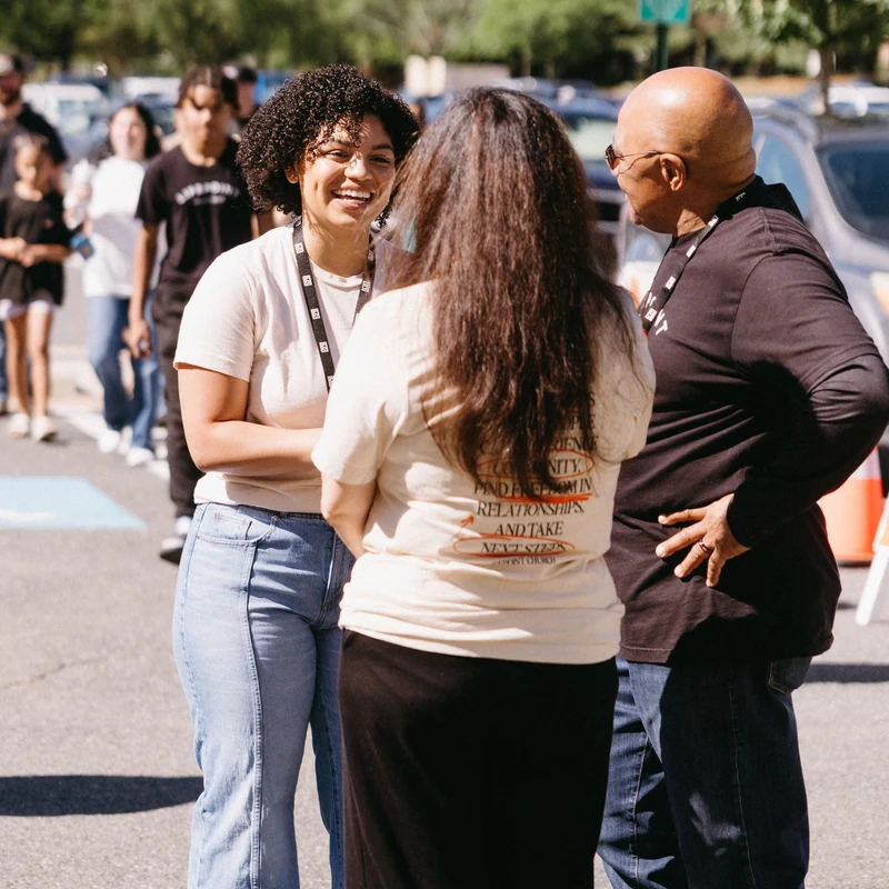 Three people stand in a sunny parking lot having a conversation. A young woman with curly hair smiles warmly while talking to a man and another woman.