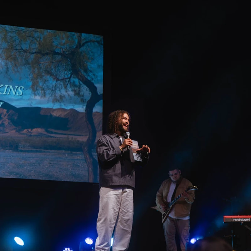 Campus Pastor Aaron Judkins speaks on a dimly lit stage with a band. Behind him, a large screen displays his name and a desert landscape.