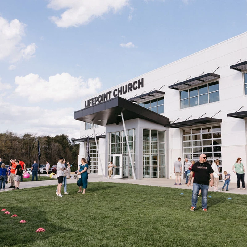 Church members gather on a lush green lawn in front of the modern Lifepoint Church building on a bright, partly cloudy day.