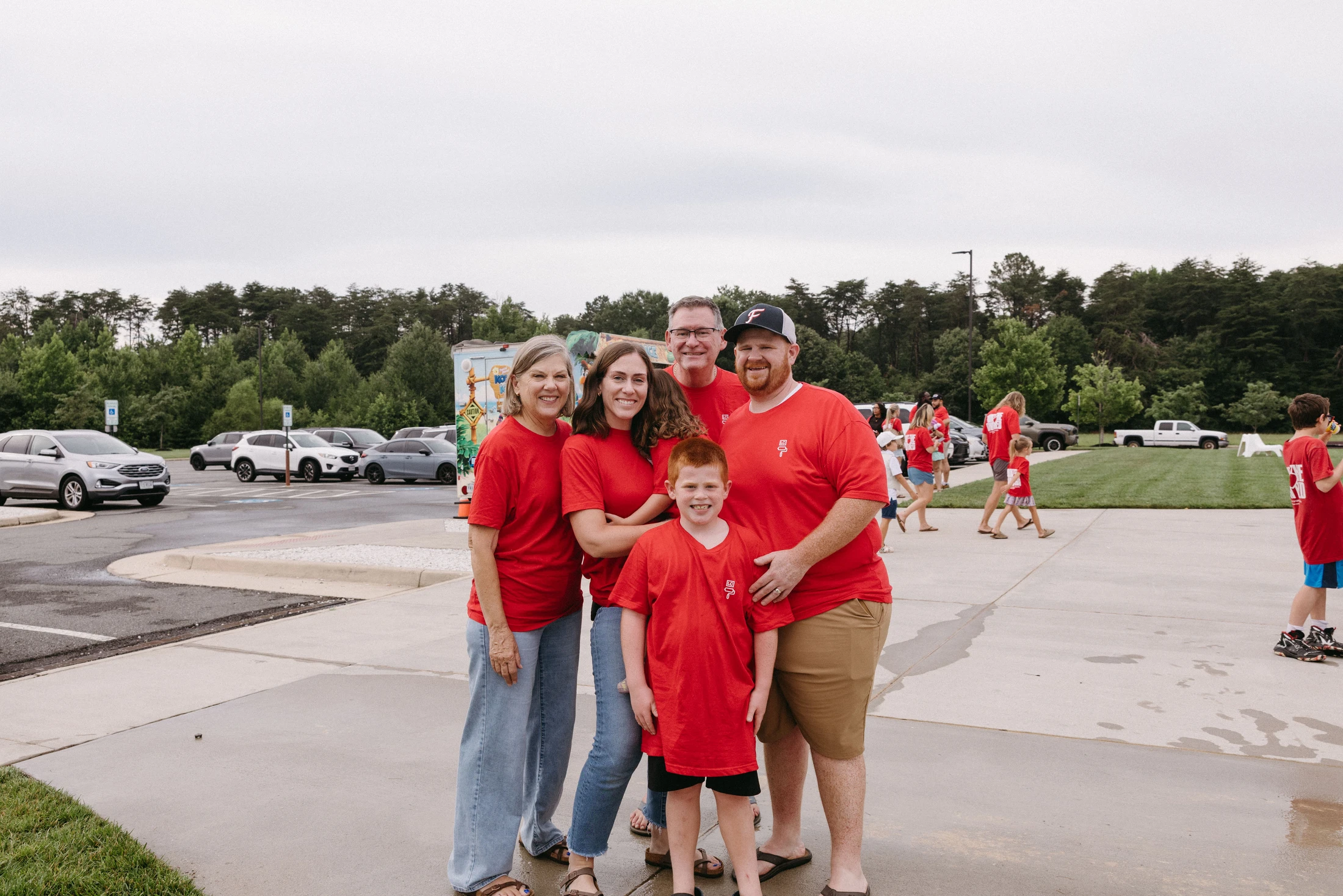 A family of five people, spanning three generations, wearing matching red t-shirts, smiles for a photo in a parking lot on an overcast day.