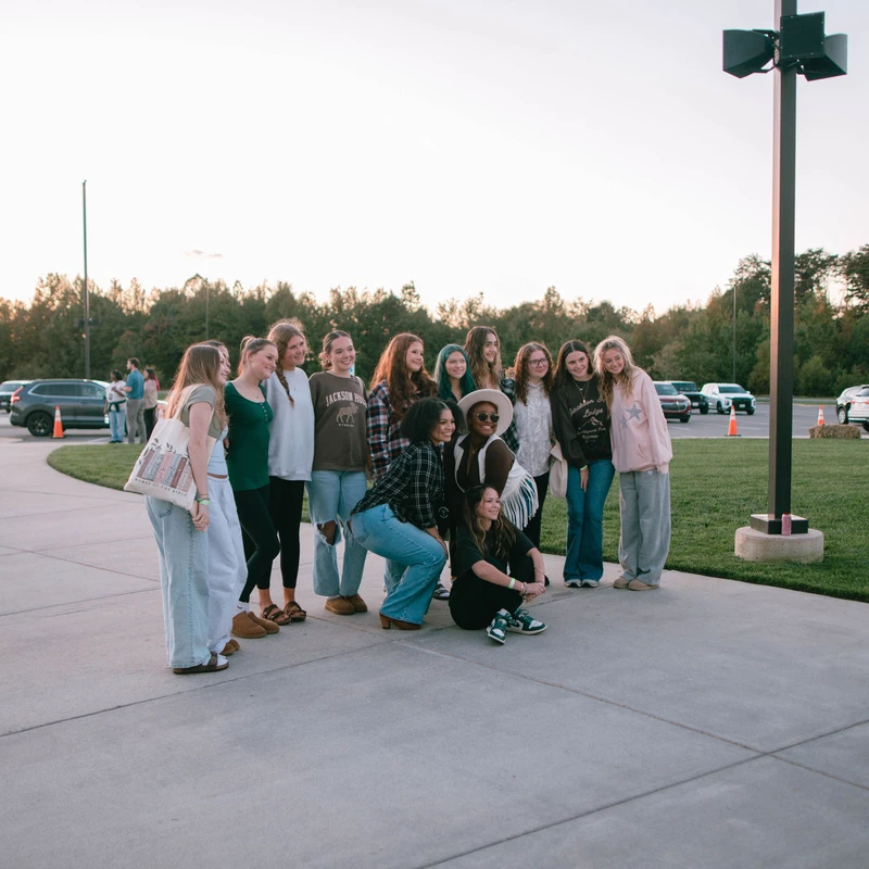 A large group of casually dressed young women smile and pose together for a picture on a sidewalk next to a lawn and parking lot during sunset.