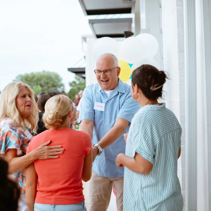 A smiling man in a blue shirt and glasses joyfully greets and shakes hands with several women at an outdoor event with balloons in the background.