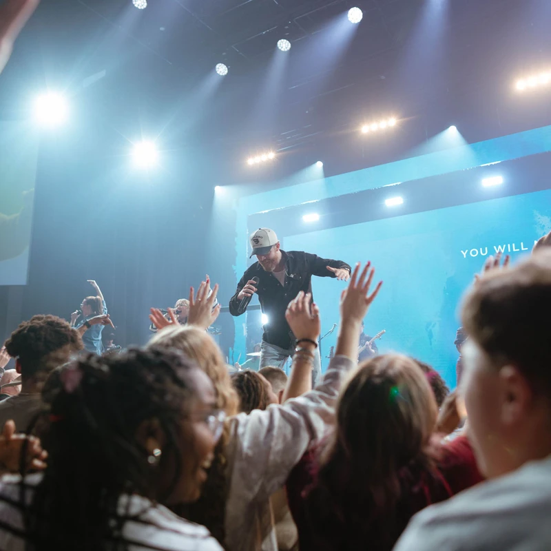 From the crowd, two male singers lead worship on a brightly lit stage as the audience raises their hands in praise during a concert or church service.