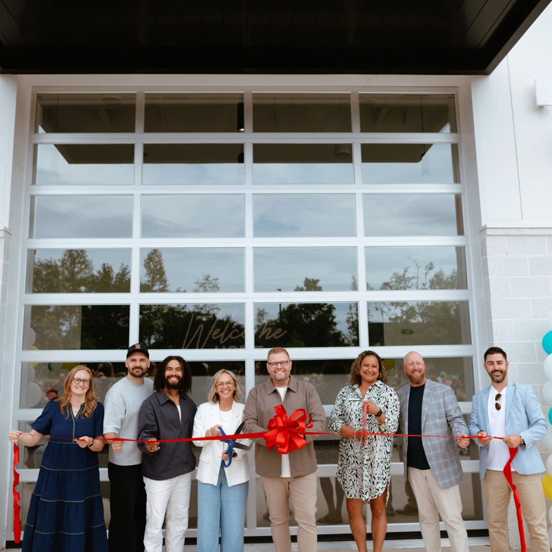 A diverse group of people smiles for the camera while holding a red ribbon during a grand opening ceremony in front of a modern building with glass doors.