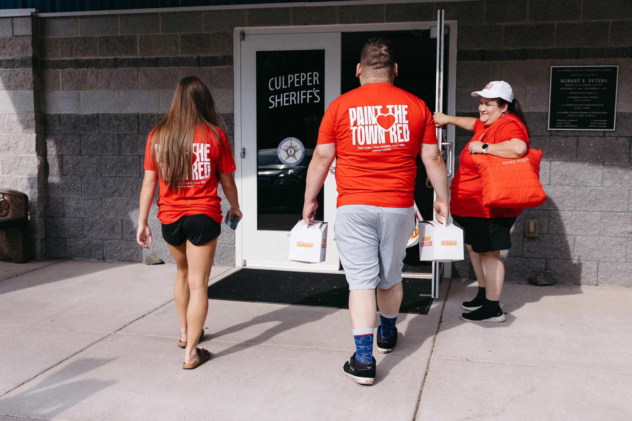 Three volunteers in matching red t-shirts bring boxes of Dunkin' coffee to the entrance of the Culpeper Sheriff's office on a sunny day.