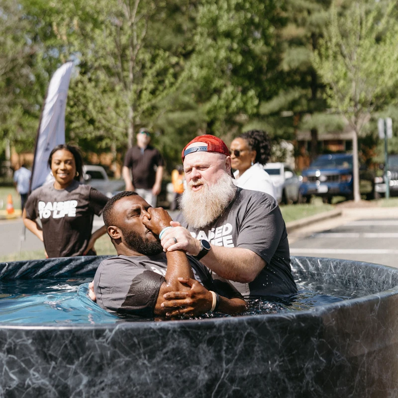 A man with a white beard baptizes another man in a portable pool outdoors, holding his nose as he lowers him into the water. Onlookers watch in the background.