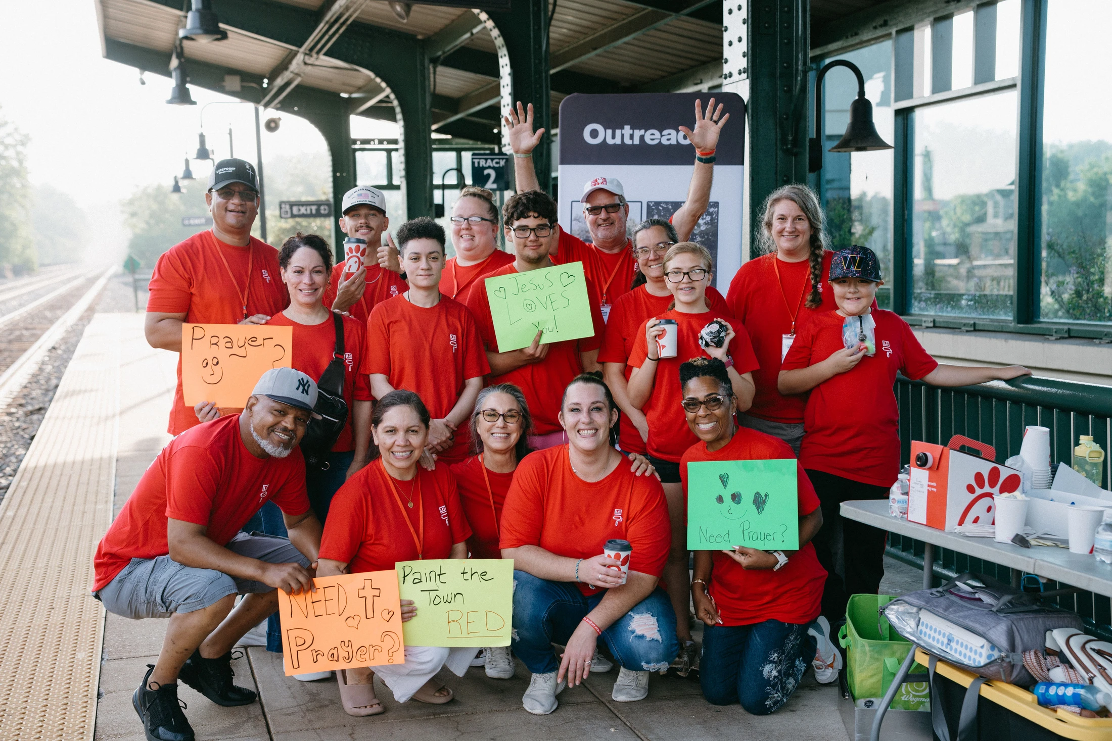 A diverse group of smiling volunteers in matching red t-shirts poses for a photo on a train platform, holding signs that offer prayer and support.