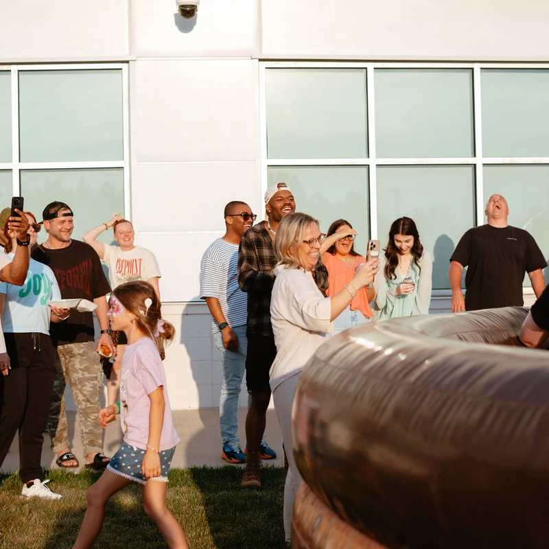 A diverse group of adults and children stand outside, laughing and taking photos as they watch an event involving a large inflatable ride.