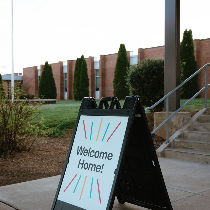 A black A-frame sign on a sidewalk reads 'Welcome Home!' outside of the Lifepoint Church building, with a lawn and trees in the background.