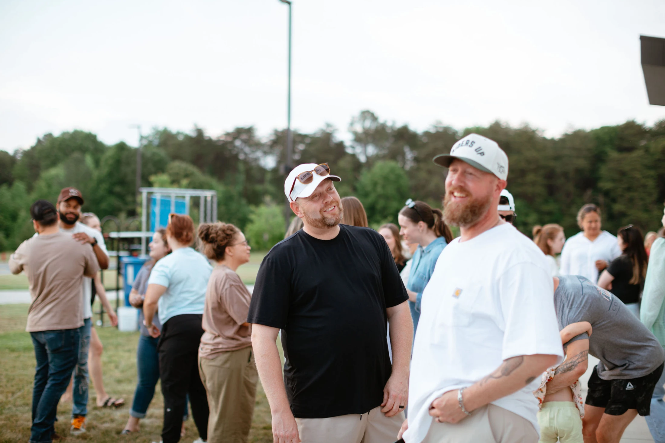 Two men wearing baseball caps smile and talk in the foreground of a large group of people mingling outdoors on a grassy lawn.