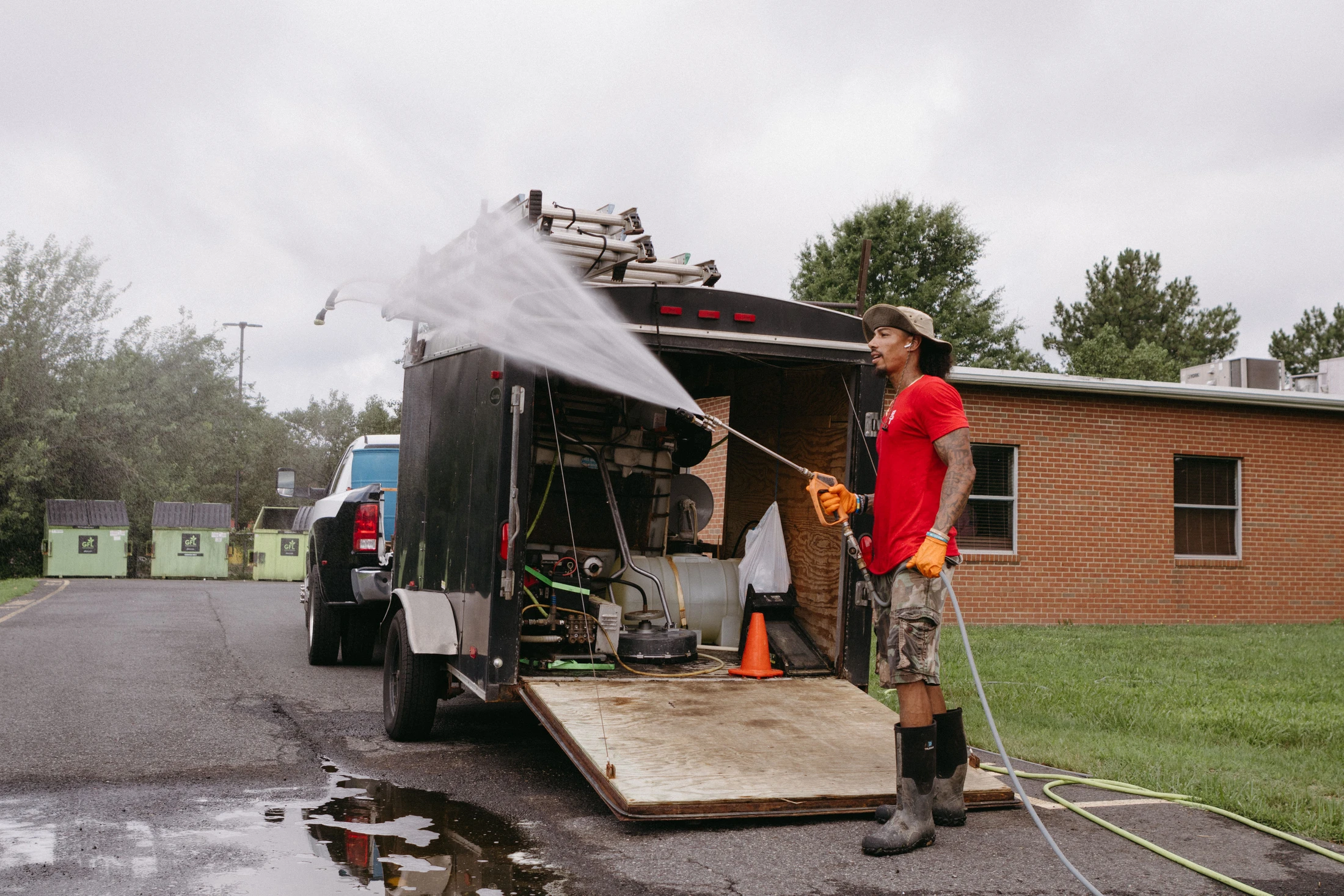 A man in a red shirt and boots stands beside a work trailer, using a high-pressure hose to spray water towards the camera on an overcast day.