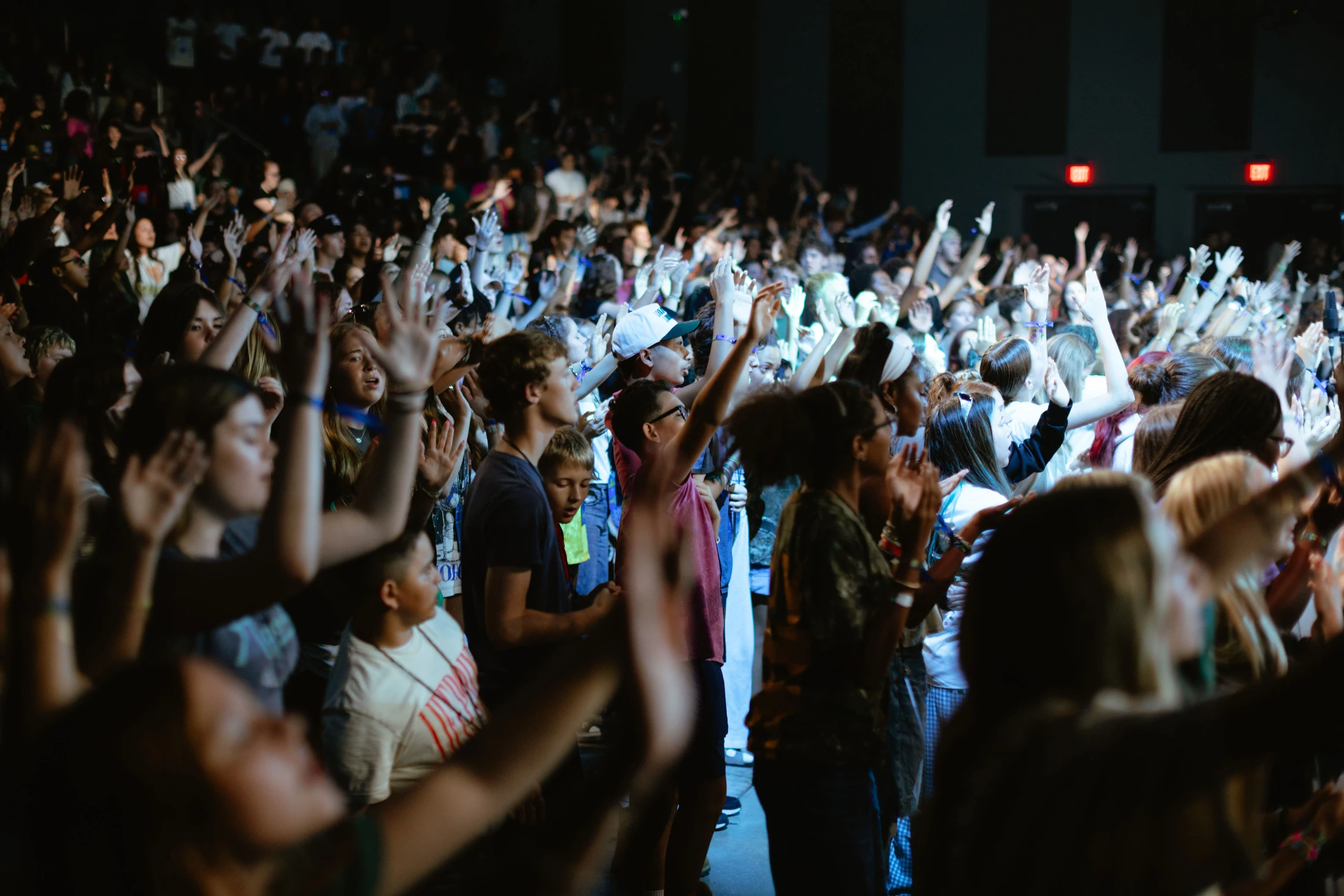 A large, diverse crowd of young people stands in a dimly lit venue, all with their hands raised in the air, participating in an event.