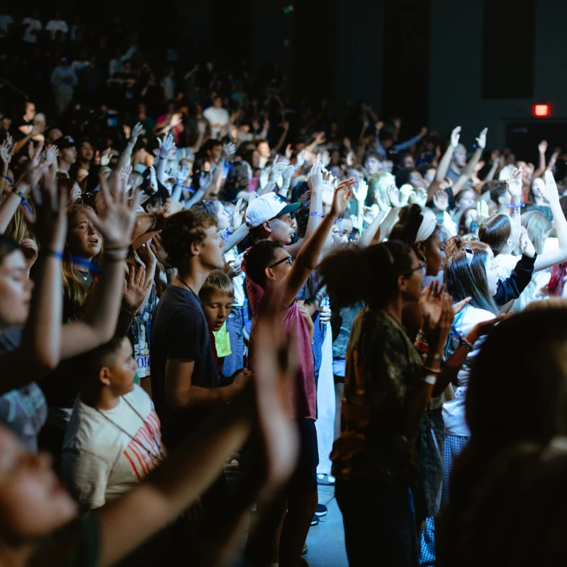 A large, diverse crowd of young people stands in a dimly lit venue, all with their hands raised in the air, participating in an event.
