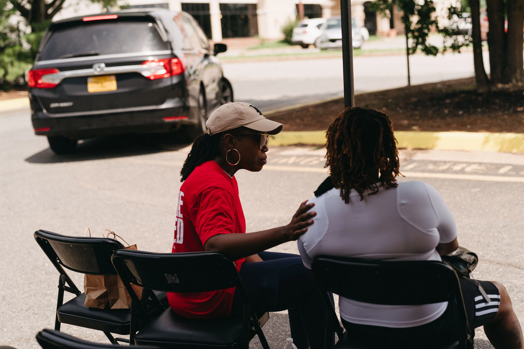 Two Black women sit in folding chairs outdoors. One woman in a red shirt and cap rests a hand on the other's shoulder as they talk in a parking lot.