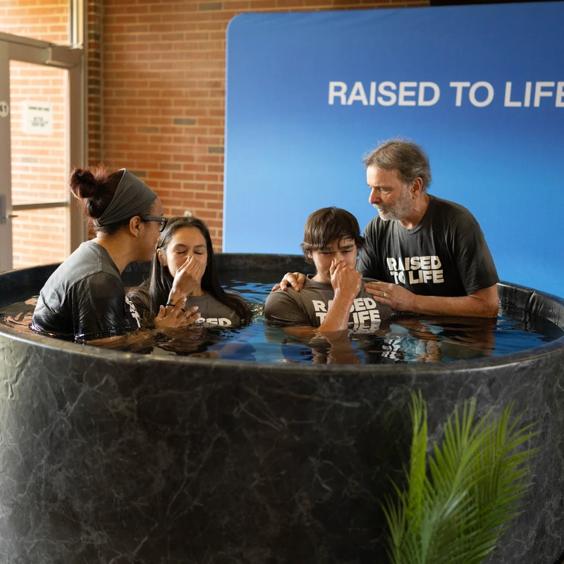 Two adults assist a young man and woman during a Christian baptism in a circular font. A blue banner behind them reads, 'RAISED TO LIFE!'