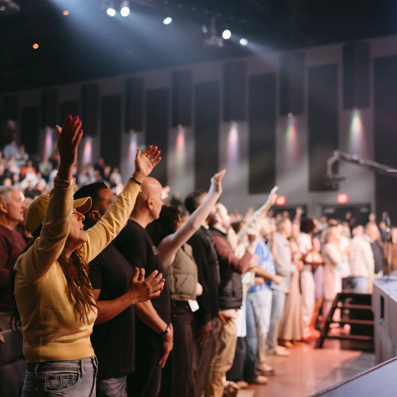 A diverse congregation stands in a dimly lit auditorium, with many raising their hands in worship. A woman in a yellow sweater is in the foreground.