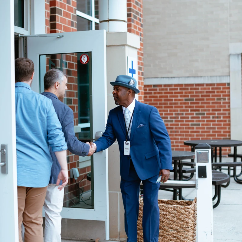 An older Black man in a blue suit and fedora smiles as he shakes hands with another man entering a brick building, welcoming him inside.