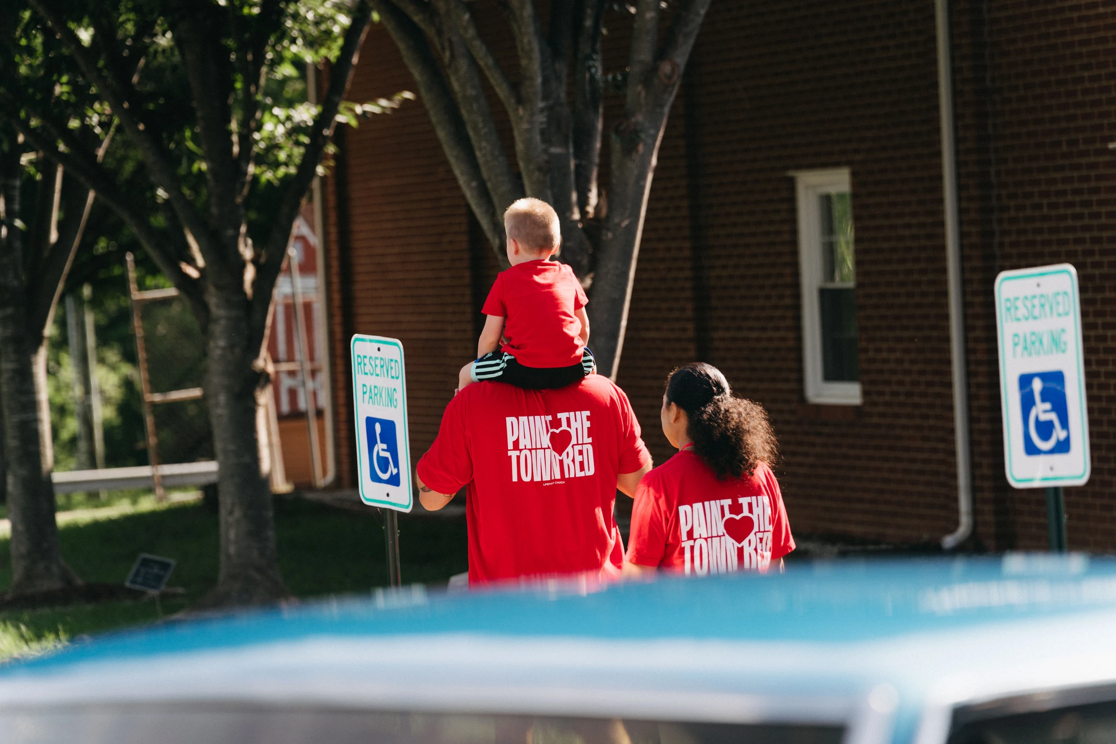 A man carries a small child on his shoulders while walking with a woman. They are all viewed from behind, wearing matching red shirts for an event.
