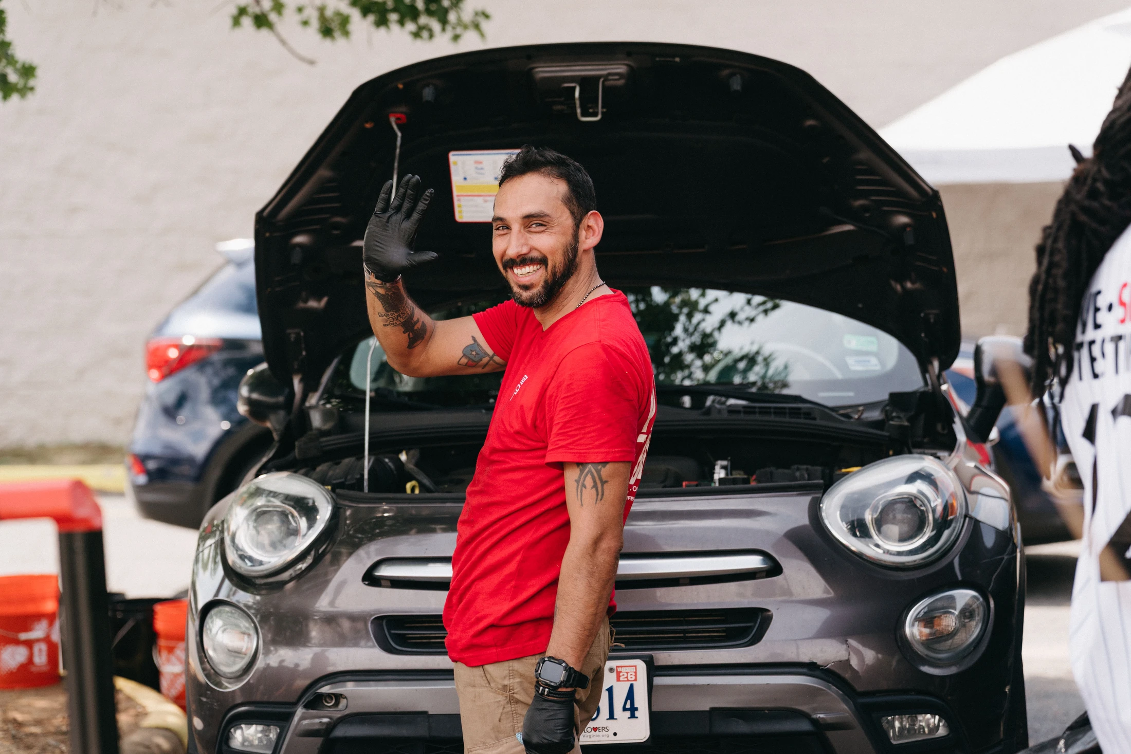 A smiling man in a red shirt and black gloves waves at the camera while standing in front of a gray car with its hood open during an outdoor event.