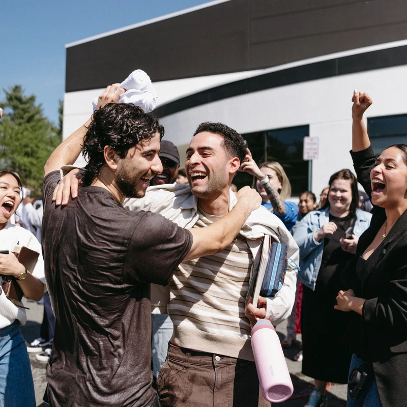 A man, soaking wet, laughs and hugs a friend, who is also laughing. They are surrounded by a cheering crowd of people outdoors on a sunny day.