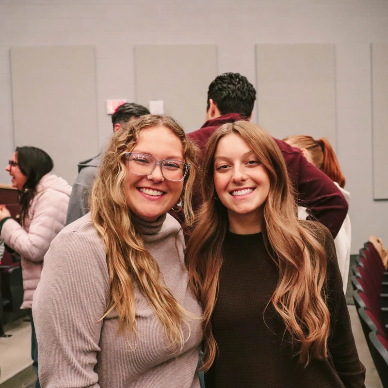 Two young women smile for the camera while standing in an auditorium. The woman on the left has blonde hair and glasses; the woman on the right has brown hair.