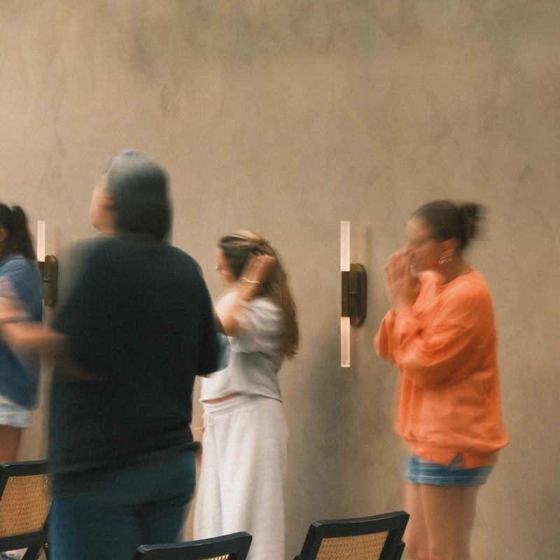 A blurry, long-exposure photograph of a group of people standing in a line, their movements creating streaks of motion against a plain wall.