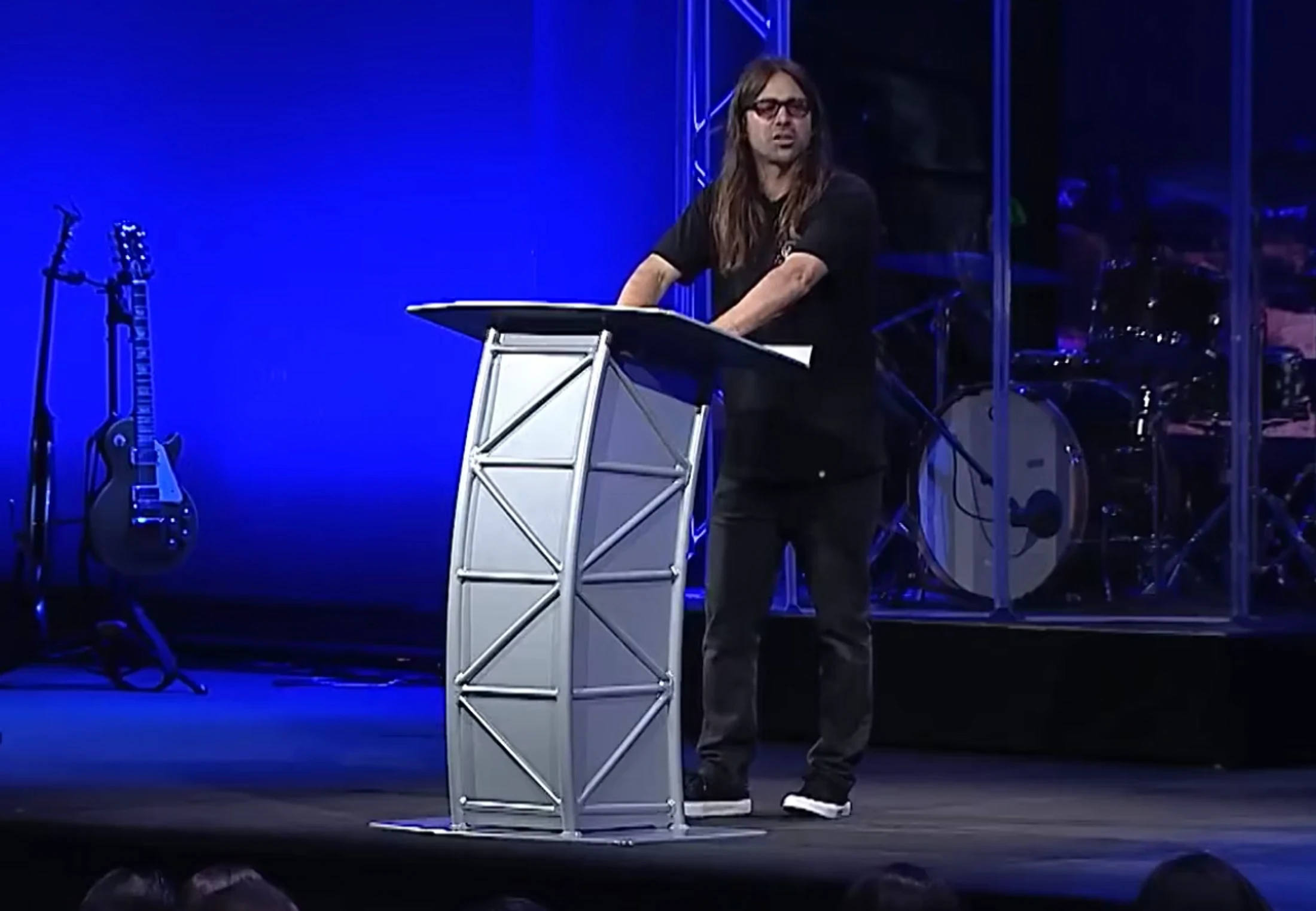 A man with long hair and glasses in a black t-shirt stands at a modern, metal lectern on a stage, with a guitar and drum set in the blue-lit background.