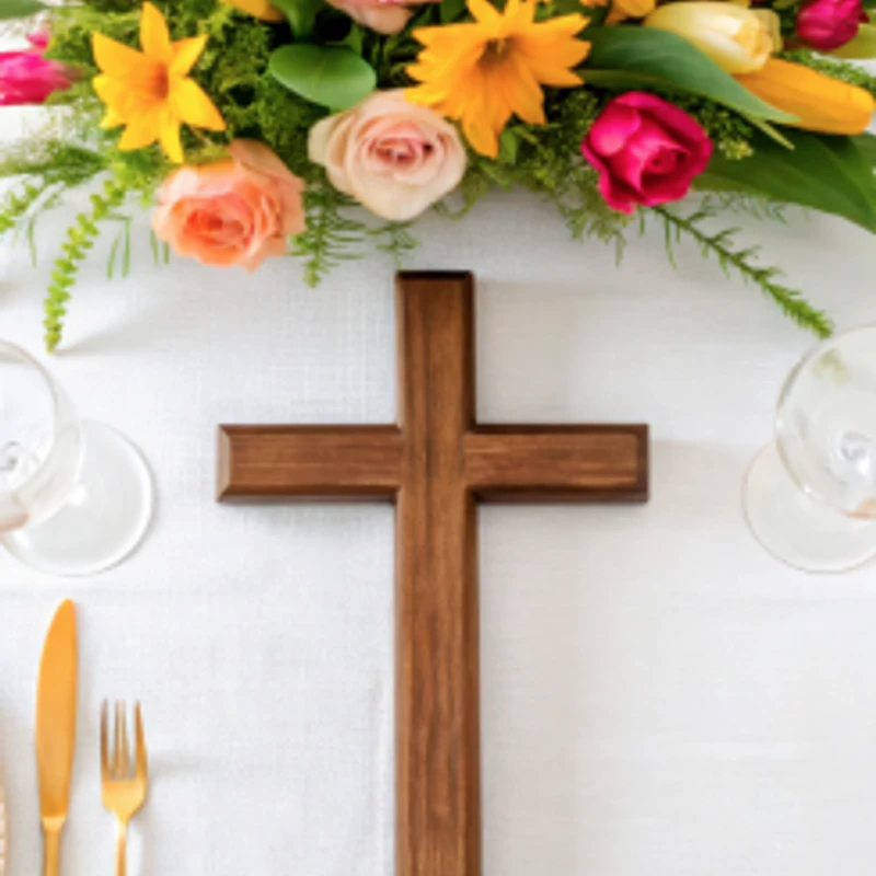 A wooden cross rests on a white tablecloth between place settings, with a colorful floral arrangement of roses and tulips at the top of the frame.