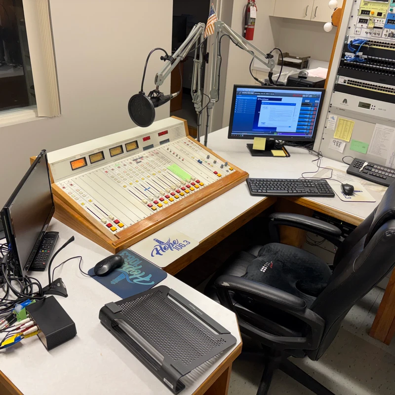 A radio station control room featuring a large audio mixing board, a microphone on a boom arm, computers, and a rack of electronic broadcasting equipment.