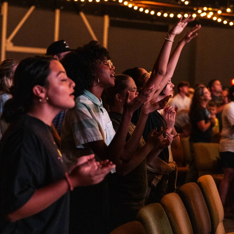 A diverse group of people stand in a warmly lit hall, singing with their hands raised in worship under strings of lights.