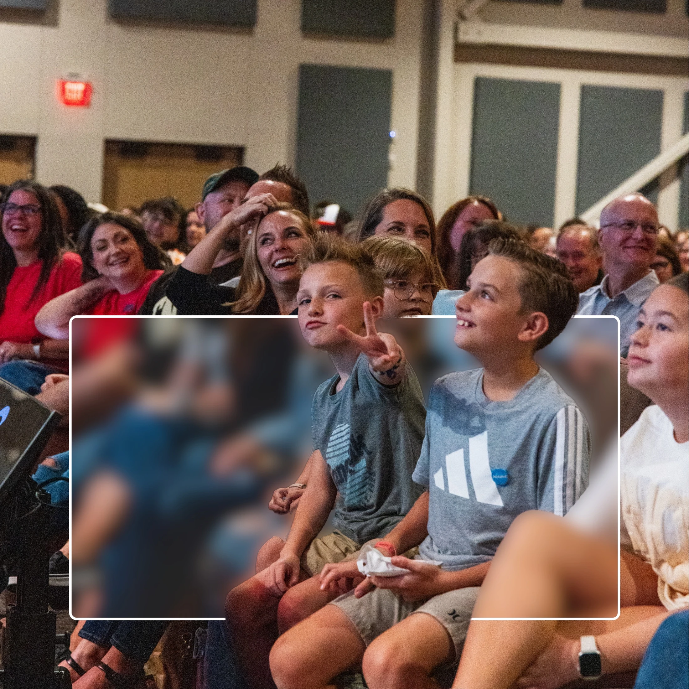 An audience of smiling adults and children watch an event. In the foreground, one boy makes a peace sign for the camera while another looks up and smiles.