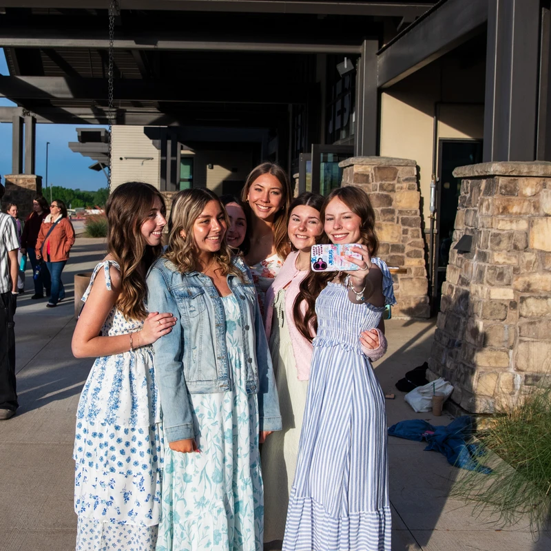 A group of six young women in summer dresses smile and pose together outside as one of them holds up a phone to take a selfie.